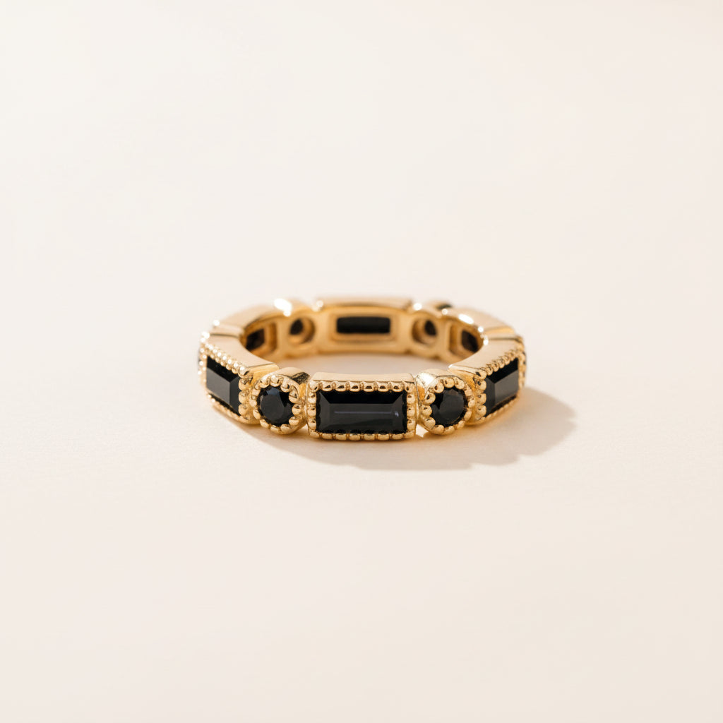 A gold band ring with rectangular black gemstones and round black accents, photographed resting on a textured stone surface with a blurred natural background.