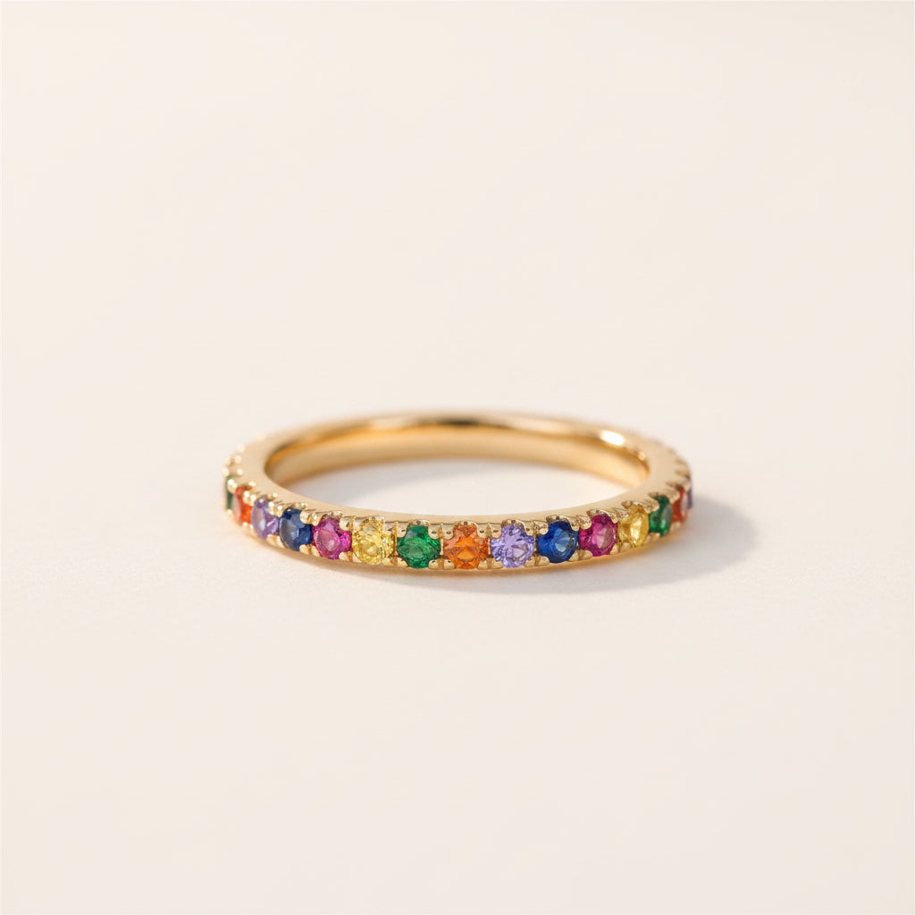 A gold eternity ring set with multiple colorful gemstones, resting on a textured stone surface with a blurred green background.