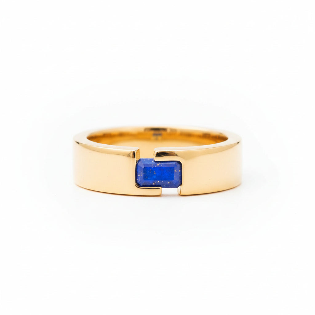 A polished gold ring with a rectangular blue sapphire stone, resting on a textured stone surface with a soft-focus green background.