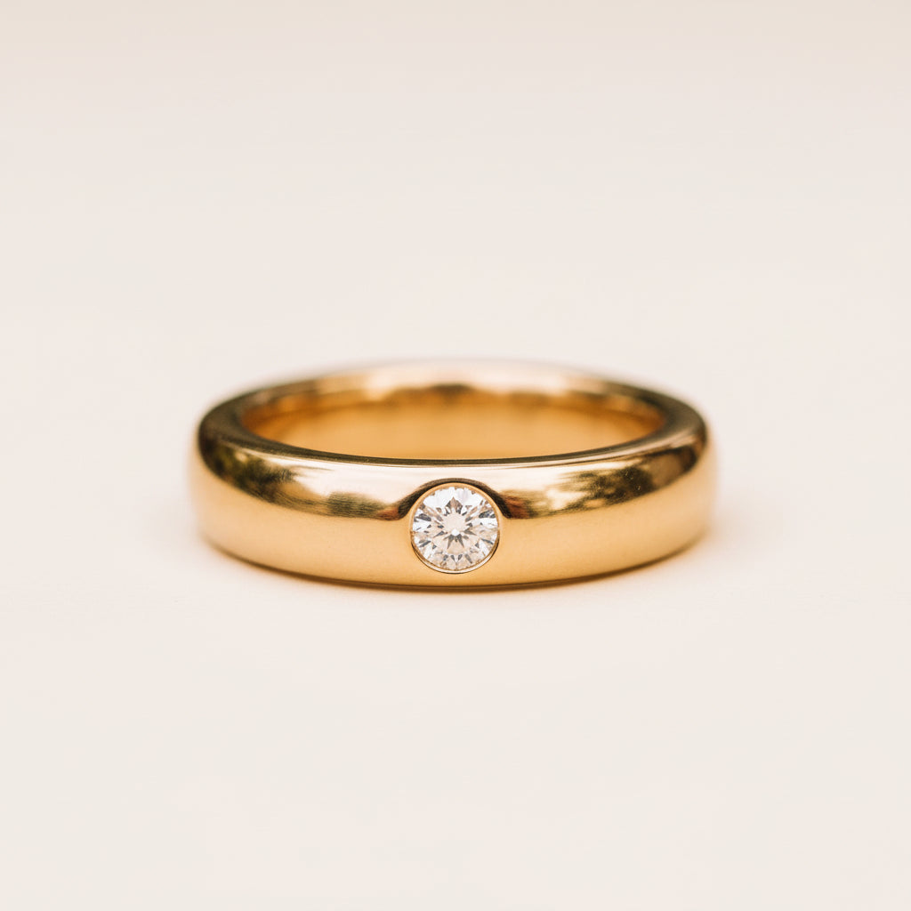 A polished yellow gold band ring with a single round-cut diamond set in a bezel setting, resting on a textured stone surface with a soft, blurred natural background.