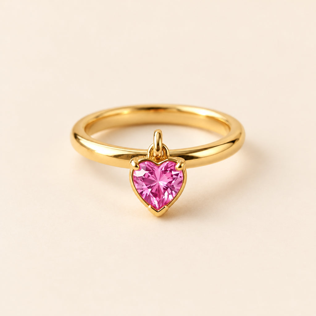 A gold ring with a heart-shaped pink gemstone, displayed on a textured beige fabric with dried flowers. The brand name 'BespokeBlingAtelier' is watermarked across the image.