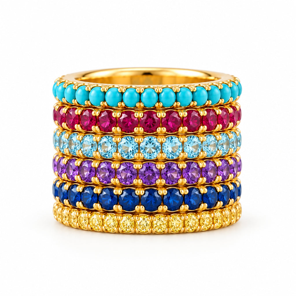 A stack of five gold rings, each set with a different row of round gemstones in turquoise, magenta, light blue, purple, and dark blue, displayed on a light marble surface.