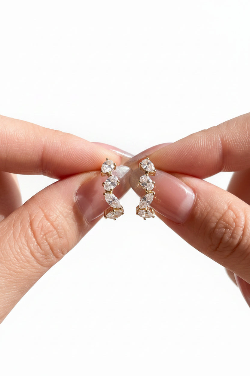 A close-up of a person's hands holding a pair of gold hoop earrings adorned with clear marquise-cut gemstones, set against a softly blurred indoor background.