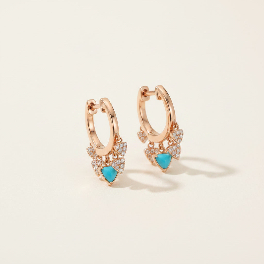 A pair of rose gold huggie hoop earrings with delicate leaf-shaped charms adorned with small clear crystals and a central turquoise blue heart, displayed on a soft white fabric background.