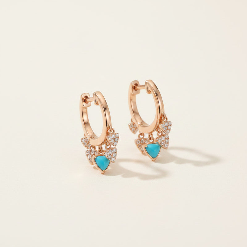 A pair of rose gold huggie hoop earrings with delicate leaf-shaped charms adorned with small clear crystals and a central turquoise blue heart, displayed on a soft white fabric background.