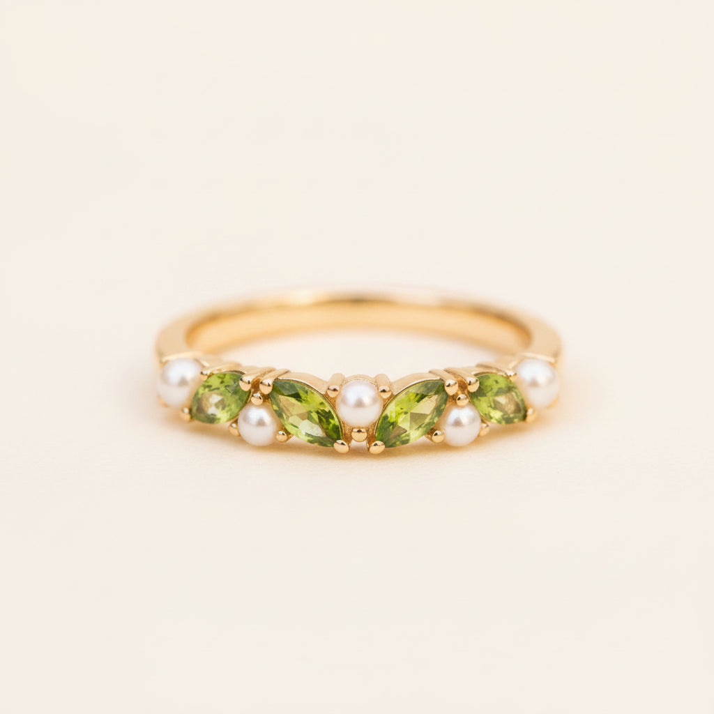 A gold ring featuring marquise-cut green peridot gemstones and white pearl accents, resting on a textured stone surface with a blurred natural background.