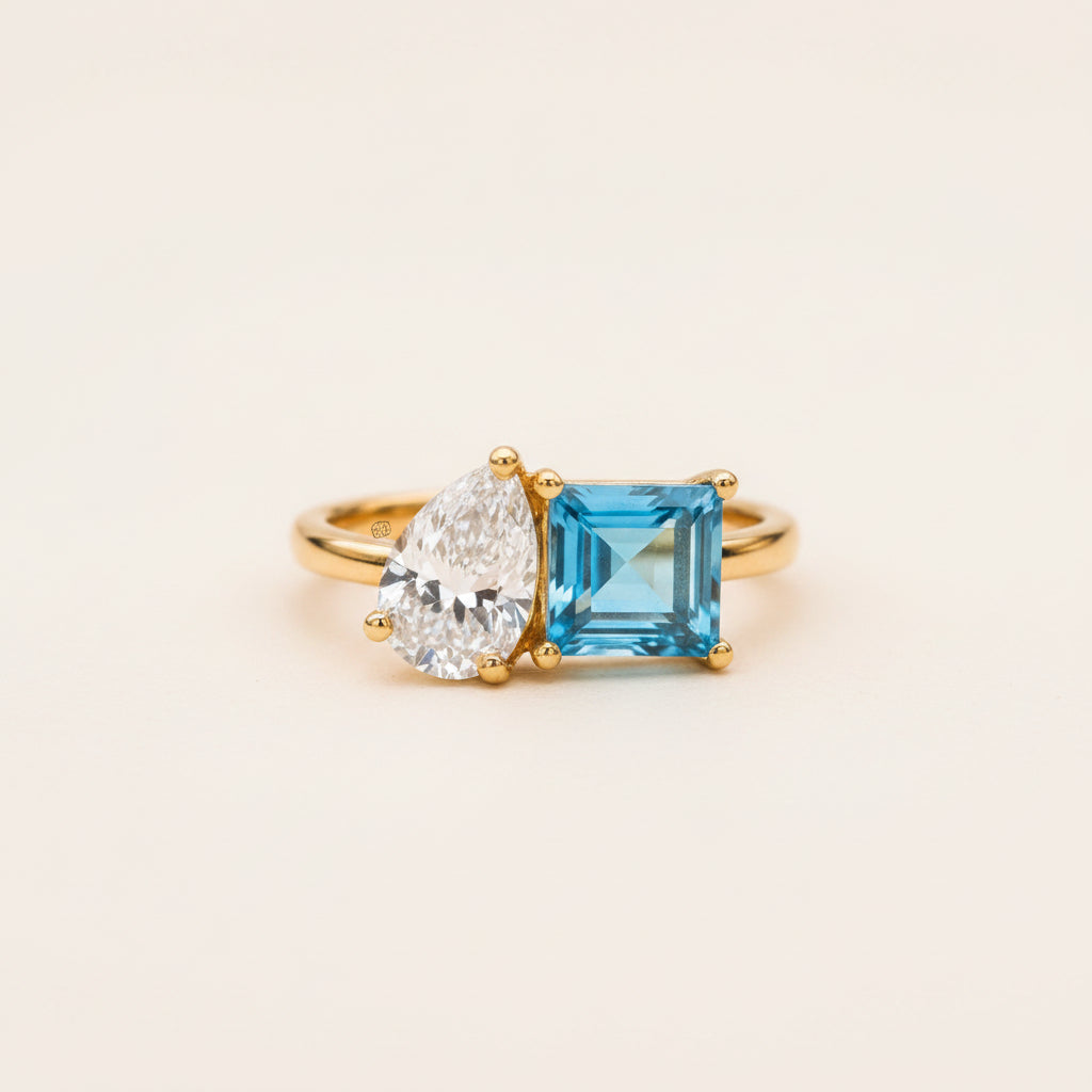 A yellow gold ring featuring a pear-cut clear diamond and a square-cut bright blue gemstone, resting on a textured stone surface with a blurred natural background.
