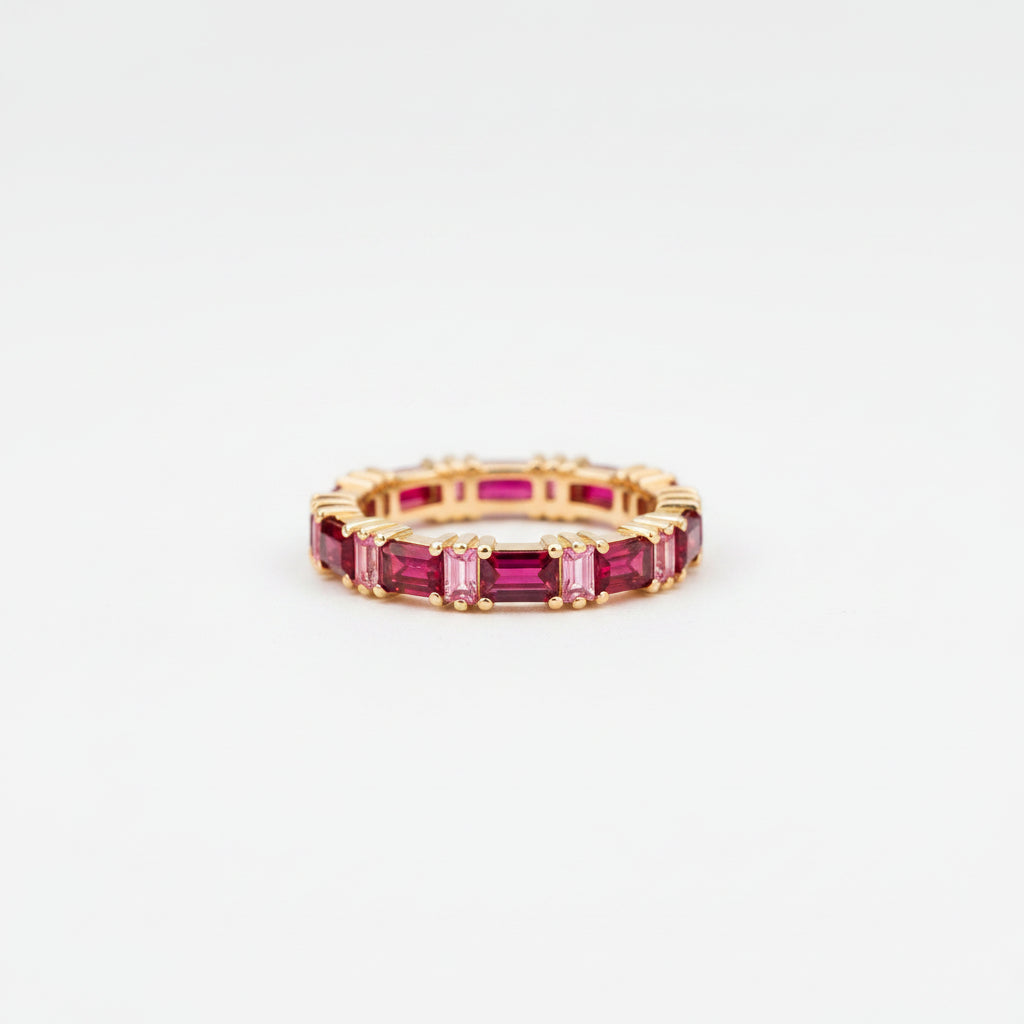 A gold eternity ring featuring baguette-cut deep pinkish-red gemstones, resting on a textured stone surface with a blurred natural background.
