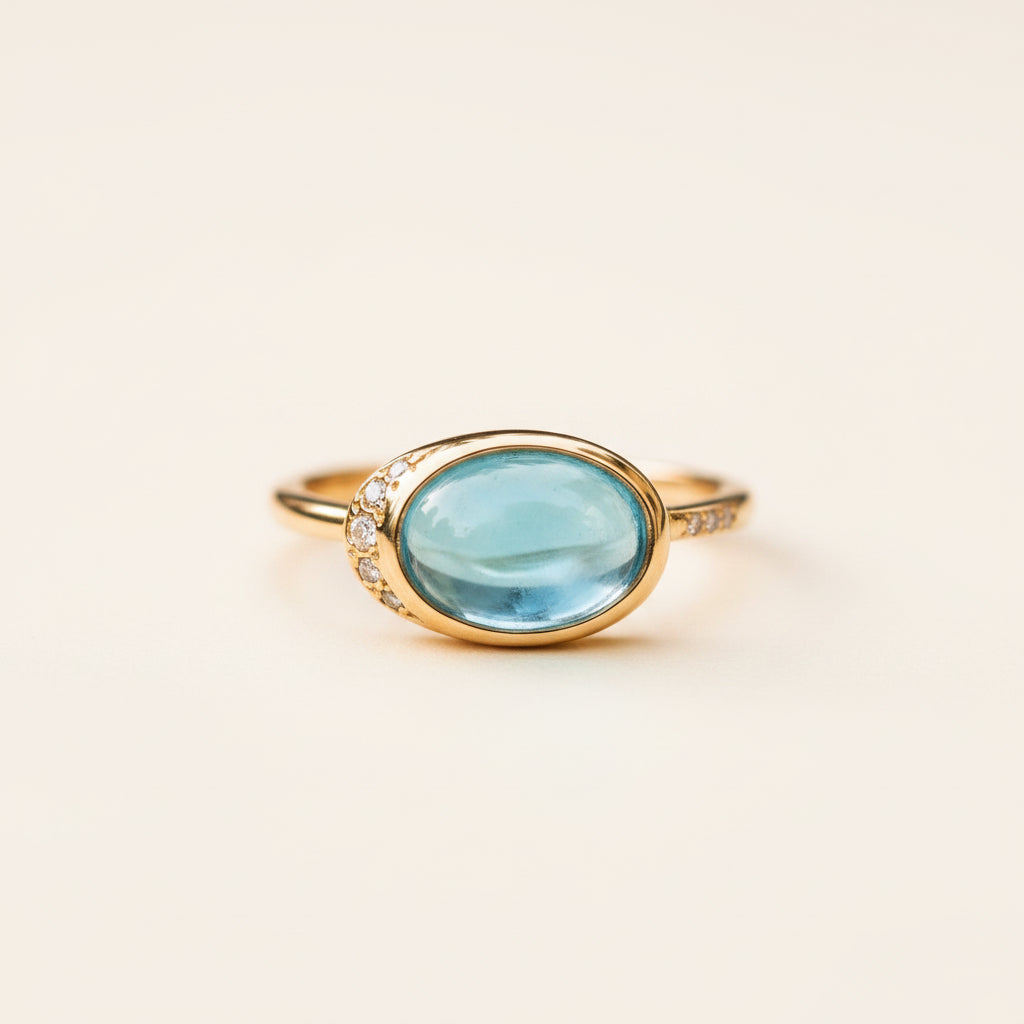 A gold ring featuring a large oval light blue gemstone, accented by small clear stones along the shoulders, resting on a textured stone surface with a blurred natural background.