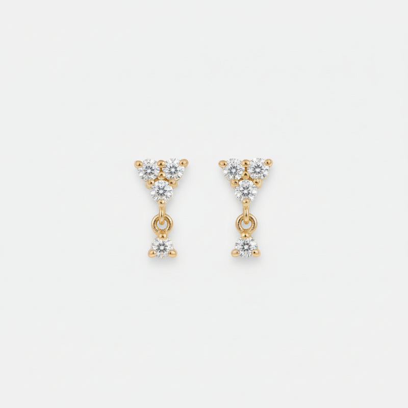 A pair of gold triangular dangle earrings with clear gemstones, resting on a draped beige satin fabric background.