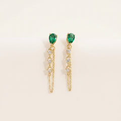 A pair of gold dangle earrings with green teardrop-shaped gemstones and clear rhinestone accents, displayed on the petals of a white rose.