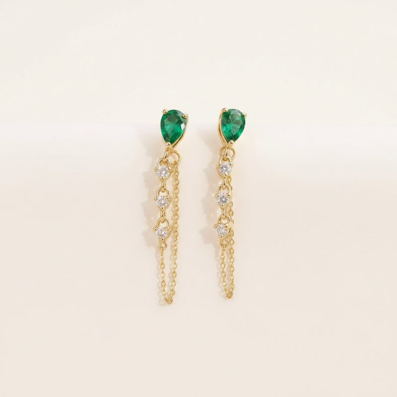 A pair of gold dangle earrings with green teardrop-shaped gemstones and clear rhinestone accents, displayed on the petals of a white rose.