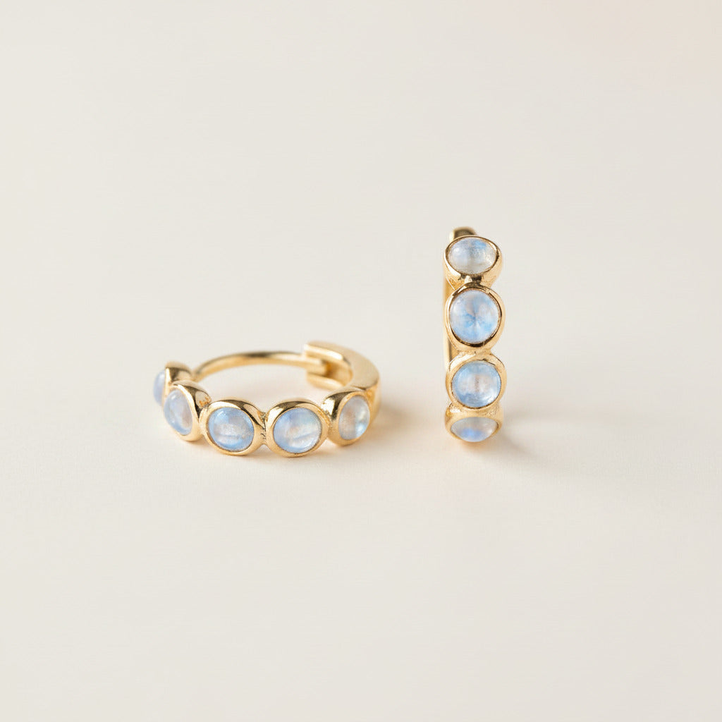 A pair of gold huggie hoop earrings with small round iridescent blue-white opal-like stones, displayed on draped cream satin fabric.