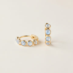 A pair of gold huggie hoop earrings with small round iridescent blue-white opal-like stones, displayed on draped cream satin fabric.