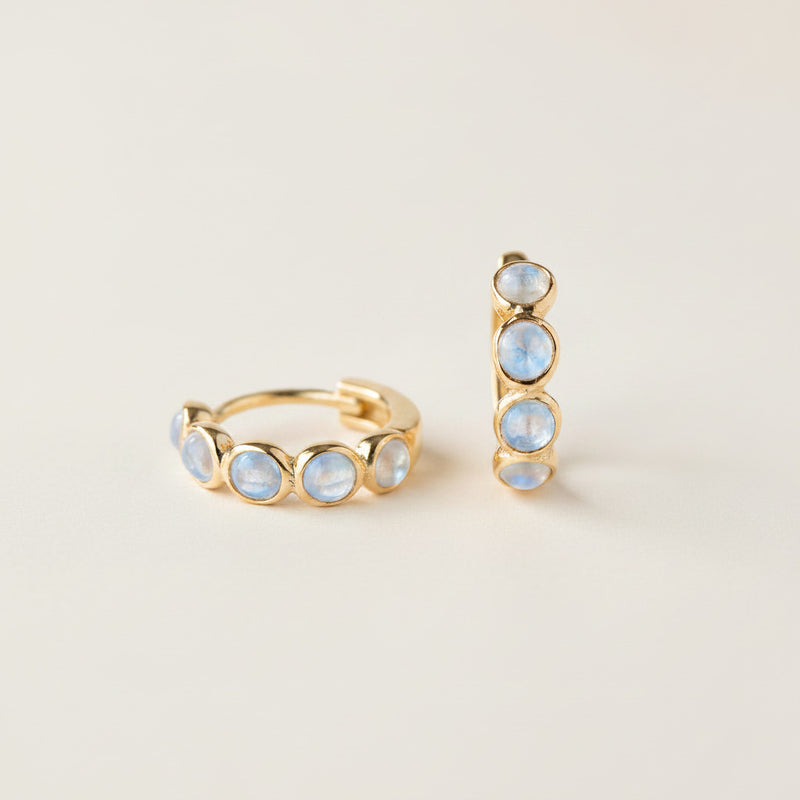 A pair of gold huggie hoop earrings with small round iridescent blue-white opal-like stones, displayed on draped cream satin fabric.