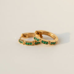 Two gold hoop rings with rows of small green stones, resting on draped white fabric.