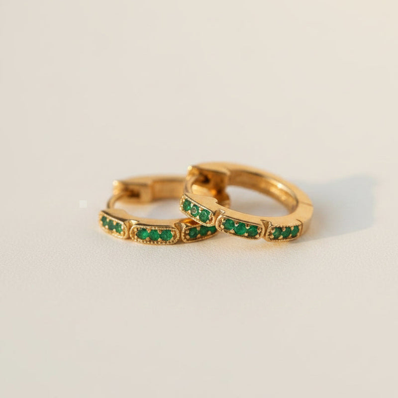 Two gold hoop rings with rows of small green stones, resting on draped white fabric.