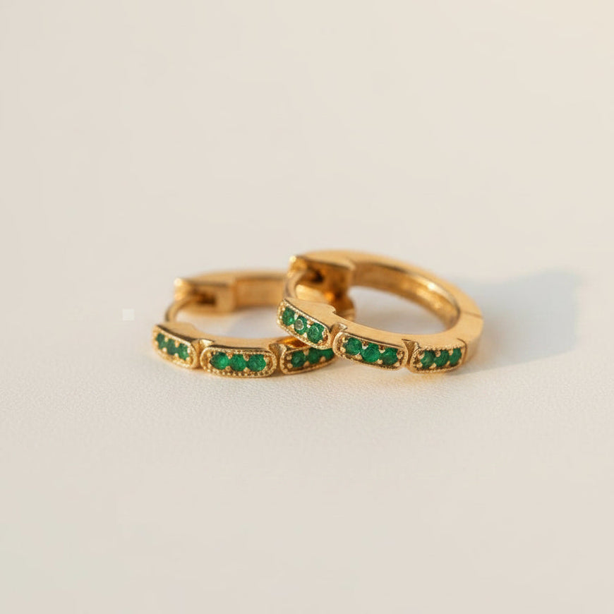 Two gold hoop rings with rows of small green stones, resting on draped white fabric.