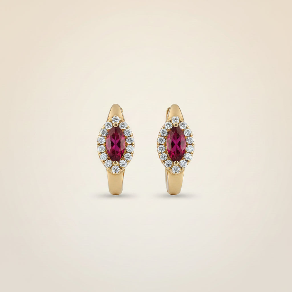 A pair of gold huggie hoop earrings with oval pinkish-red gemstones surrounded by small clear stones, displayed on a soft beige fabric background.