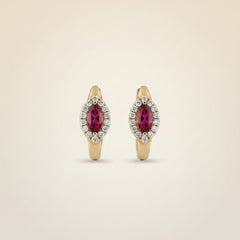A pair of gold huggie hoop earrings with oval pinkish-red gemstones surrounded by small clear stones, displayed on a soft beige fabric background.