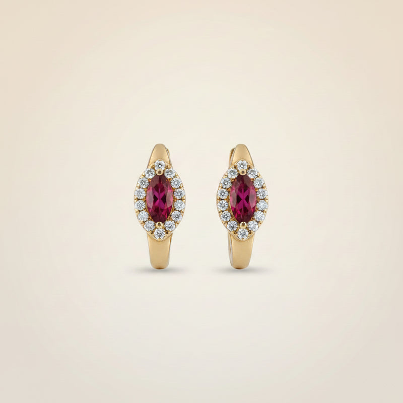 A pair of gold huggie hoop earrings with oval pinkish-red gemstones surrounded by small clear stones, displayed on a soft beige fabric background.