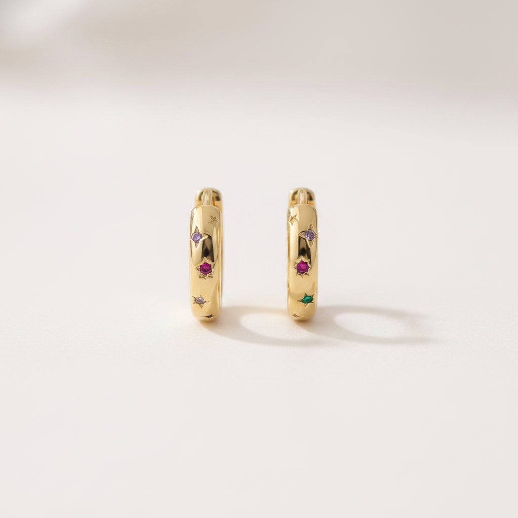 A pair of gold huggie hoop earrings with small colorful gemstone accents, displayed on a white marble cylinder against a soft, blurred light background.