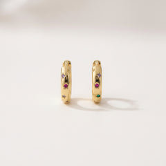 A pair of gold huggie hoop earrings with small colorful gemstone accents, displayed on a white marble cylinder against a soft, blurred light background.
