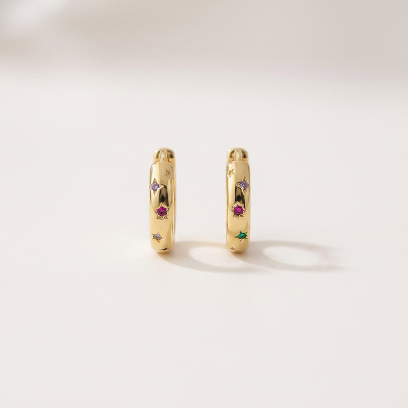 A pair of gold huggie hoop earrings with small colorful gemstone accents, displayed on a white marble cylinder against a soft, blurred light background.