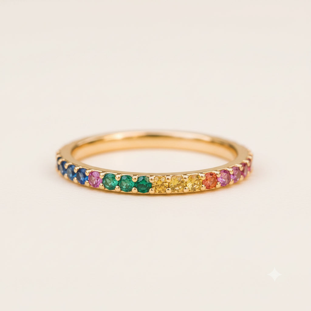 A gold eternity ring set with rainbow-colored gemstones, resting on a textured stone surface with a blurred natural background.