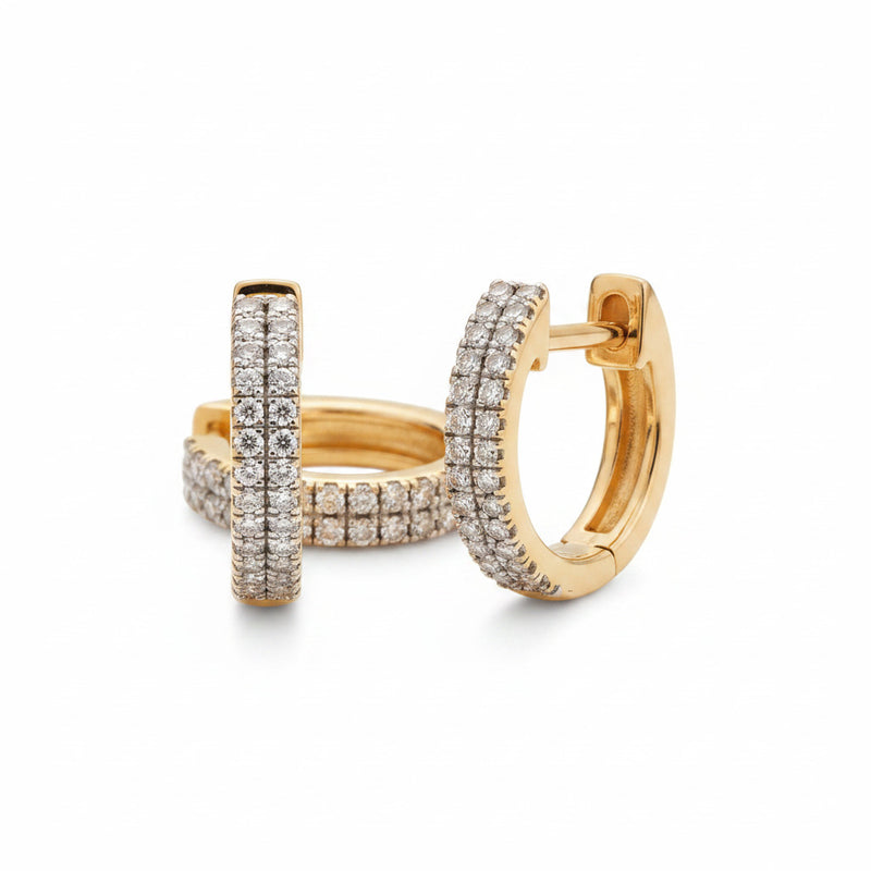 A pair of gold huggie hoop earrings paved with small clear stones, resting on a textured beige fabric background.
