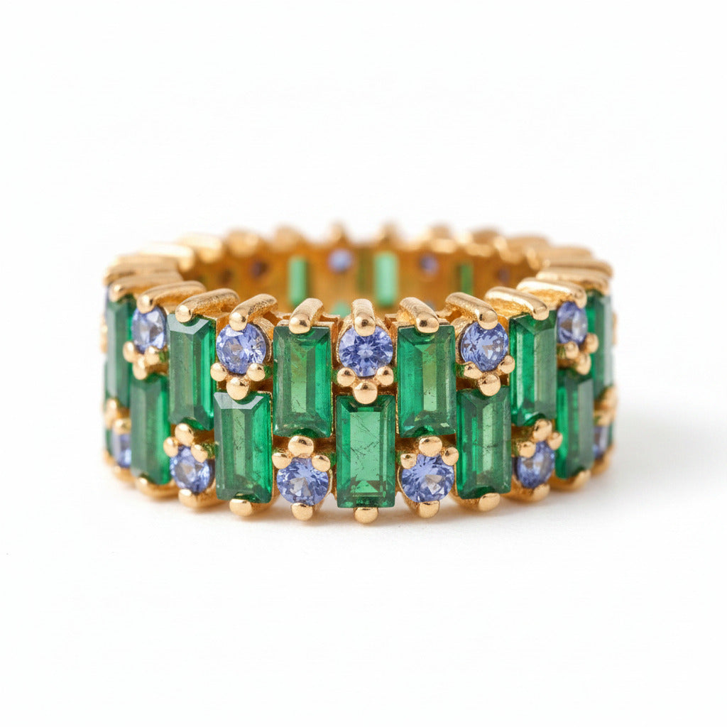 A gold eternity ring featuring emerald-cut green gemstones and round blue gemstones, displayed on a soft beige fabric background.