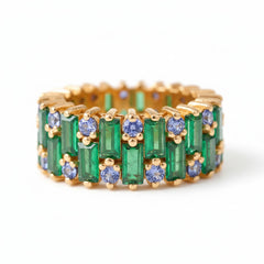 A gold eternity ring featuring emerald-cut green gemstones and round blue gemstones, displayed on a soft beige fabric background.