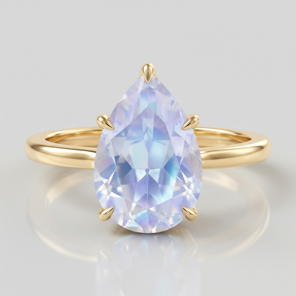 A pear-cut iridescent light blue gemstone ring set in a polished yellow gold band, resting on a white and gray marble surface.