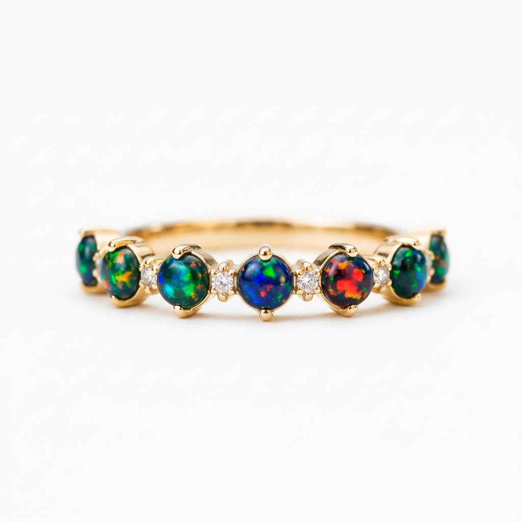 A gold band ring featuring seven round opal gemstones in vibrant green, blue, red, and black flashes, displayed on a rustic stone surface with a blurred natural background.