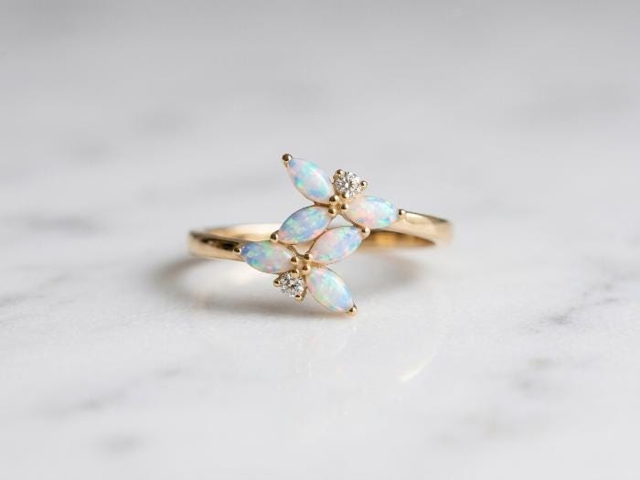 A gold ring featuring four marquise-cut iridescent opal stones and two small clear stones, displayed on a light marble surface.