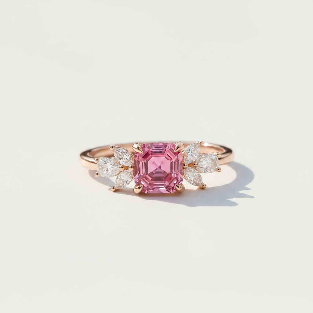 A rose gold ring featuring a large emerald-cut pink center stone and marquise-cut side stones, resting on a textured stone surface with soft leaf shadows.