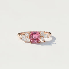 A rose gold ring featuring a large emerald-cut pink center stone and marquise-cut side stones, resting on a textured stone surface with soft leaf shadows.