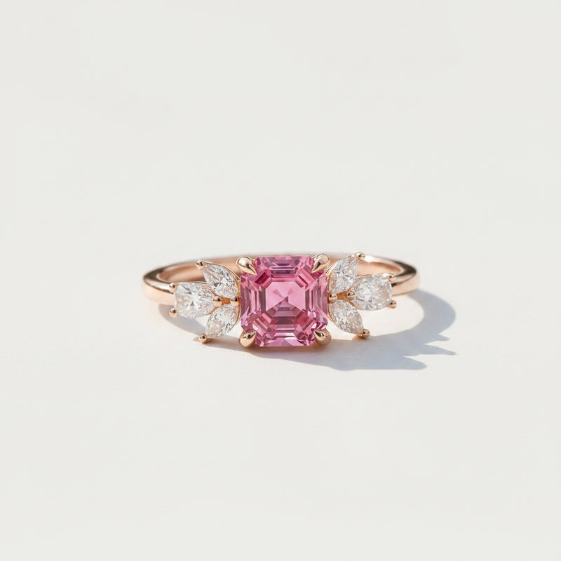 A rose gold ring featuring a large emerald-cut pink center stone and marquise-cut side stones, resting on a textured stone surface with soft leaf shadows.