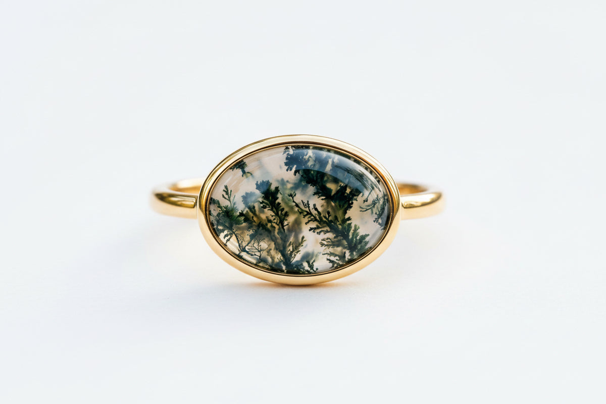 A gold band ring with an oval cabochon stone featuring a detailed botanical design of dark green pine branches and light blue-gray sky, resting on a textured stone surface with a blurred green background.