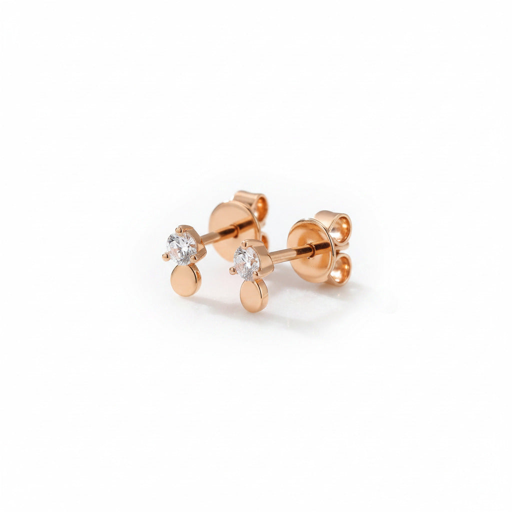 A pair of rose gold stud earrings with a small round clear gemstone and a flat circular drop, displayed on a white marble block.