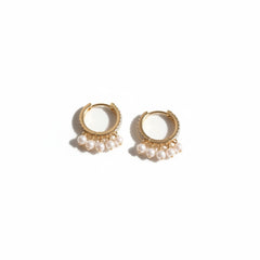 A pair of gold huggie hoop earrings with dangling pearl clusters, displayed on a white marble block with soft shadows.
