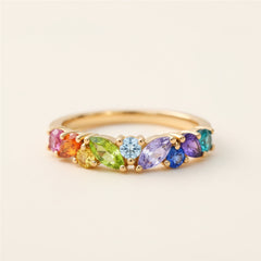 A gold ring featuring a row of colorful marquise and round gemstones in pink, orange, yellow, green, light blue, purple, and teal, resting on a textured stone surface with a blurred green background.