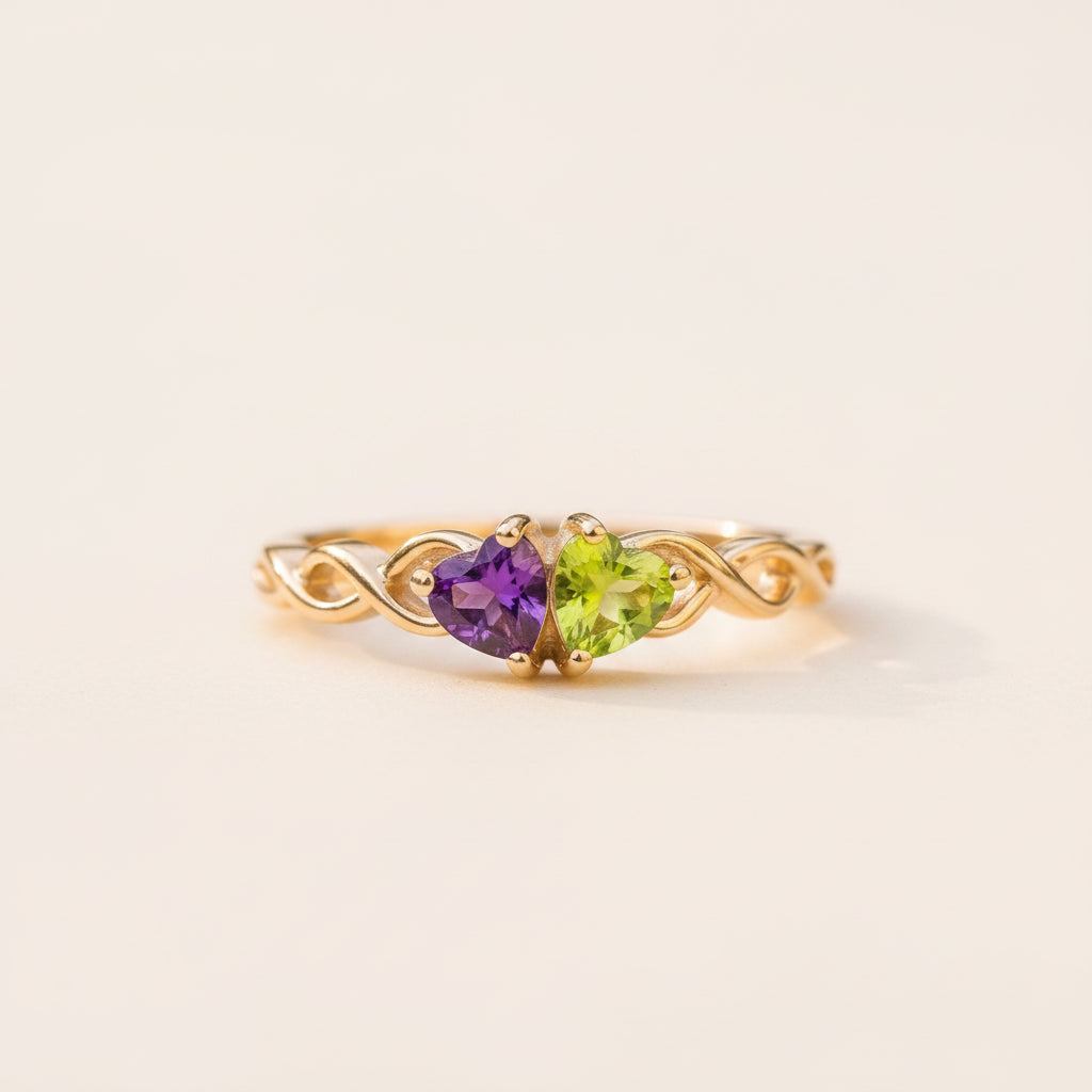 A gold ring with two heart-shaped gemstones, one purple amethyst and one green peridot, resting on a textured stone surface with a blurred natural background.