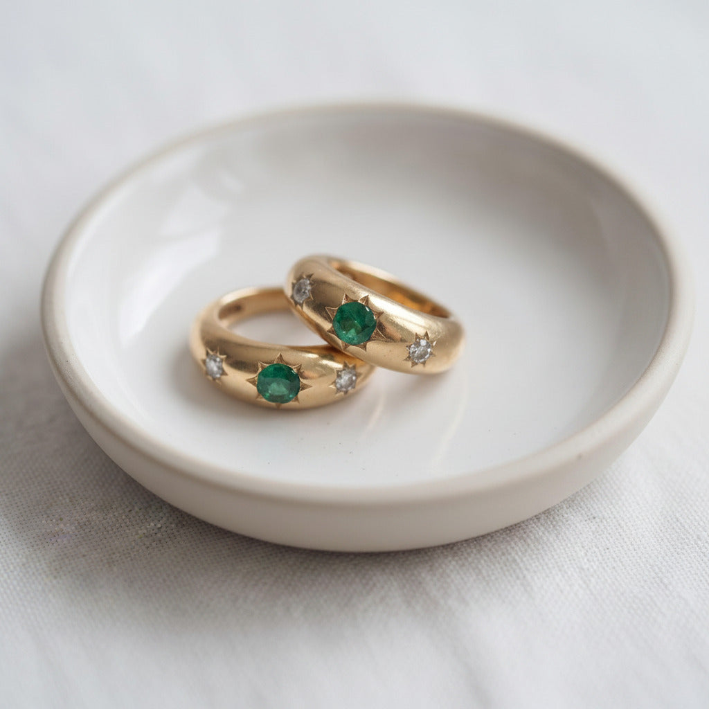 Two gold rings with green emeralds and small clear stones, displayed on a small white ceramic dish over a light fabric surface.