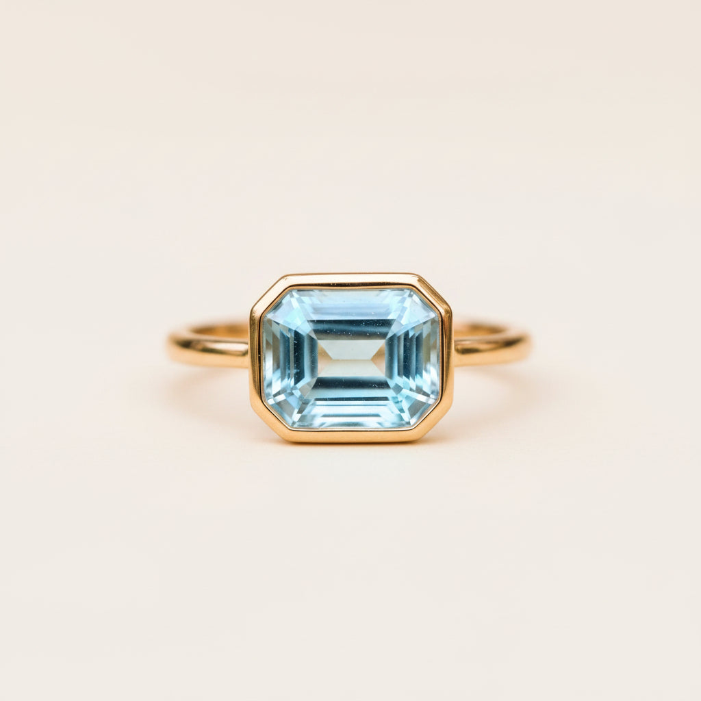 A gold ring with a large, faceted light blue topaz gemstone, resting on a textured stone surface with a blurred natural background.