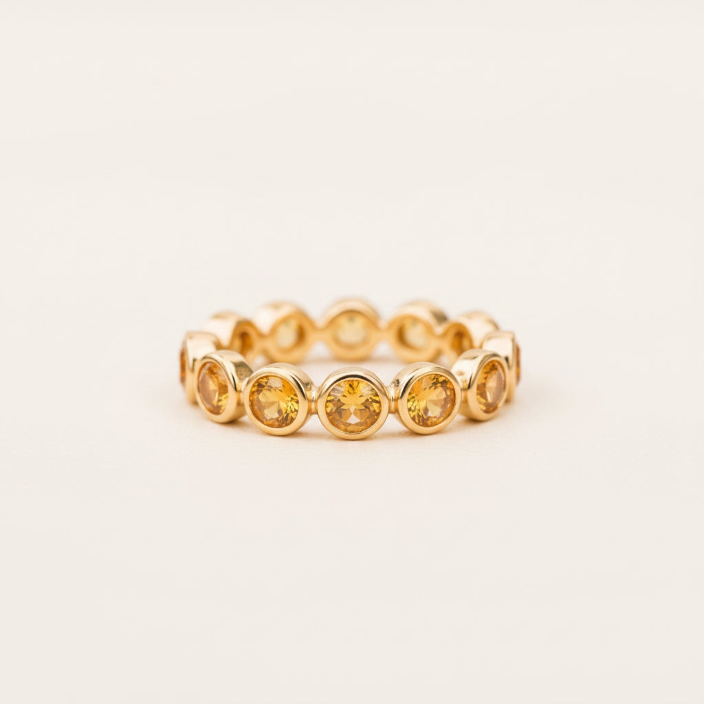 A gold-toned stretch band ring featuring a row of round, faceted amber-yellow gemstones, resting on a textured stone surface with a blurred green background.
