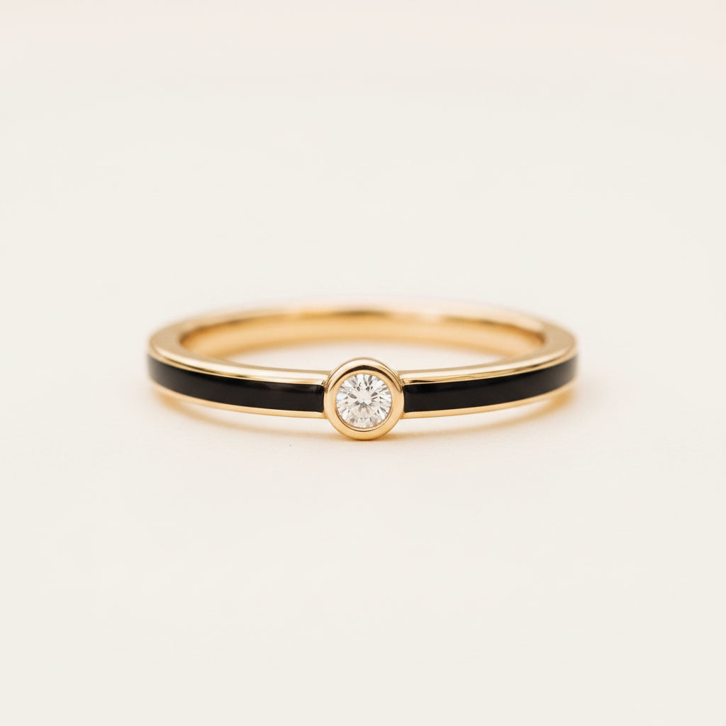 A gold ring with a central round-cut diamond and a black inlay stripe, resting on a textured stone surface with a soft-focus green bokeh background.