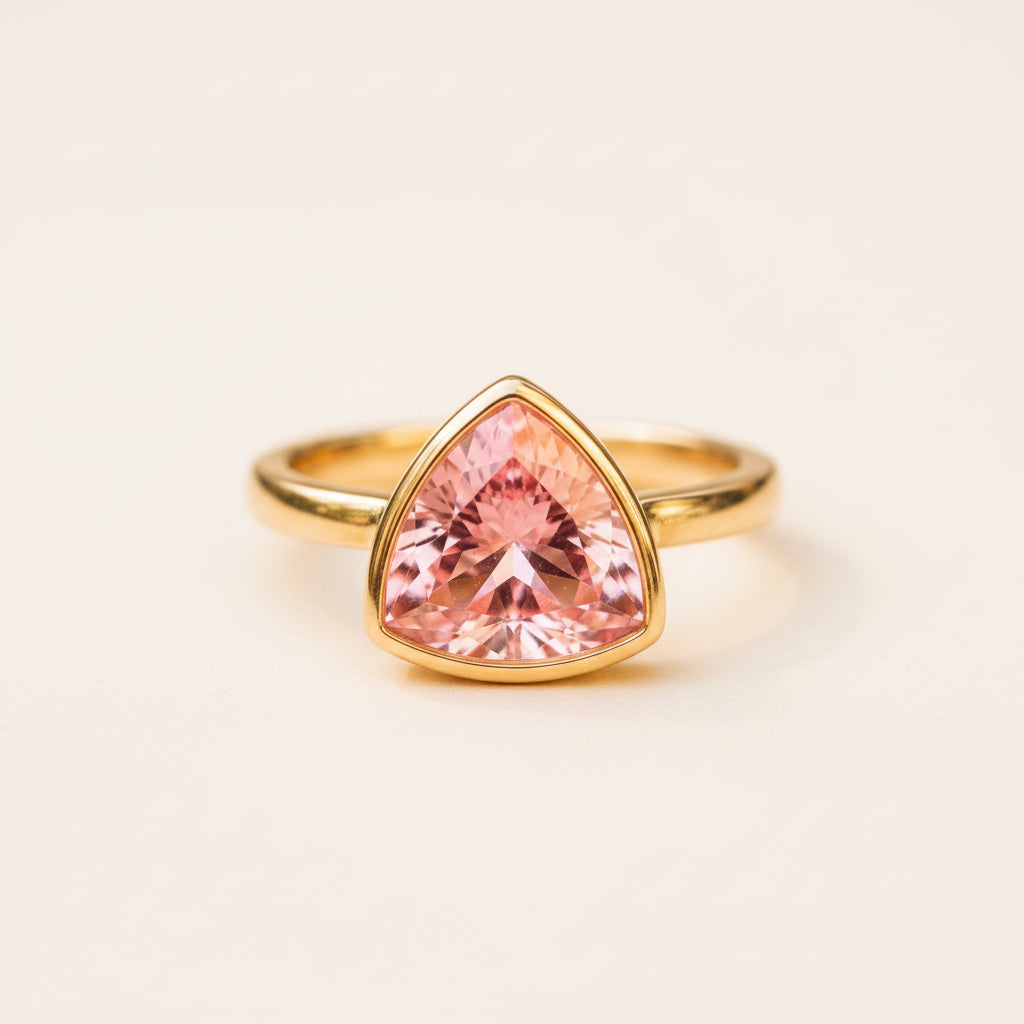 A gold ring featuring a large, faceted pink triangular gemstone, resting on a textured stone surface with a soft, blurred background.