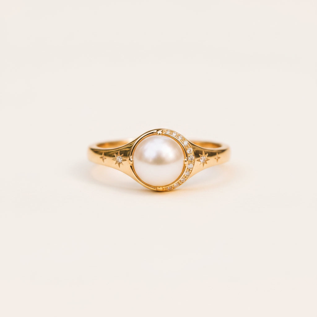 A gold ring featuring a large round white pearl in the center, surrounded by small clear stones and star motifs, resting on a textured stone surface with a blurred green background.