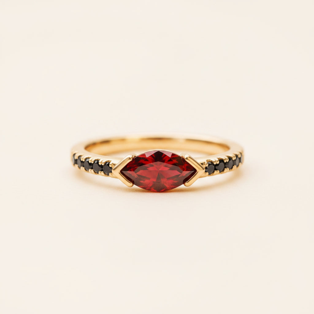 A gold ring with a marquise-cut red gemstone and black pavé-set accents on the band, resting on a textured stone surface with a blurred natural background.